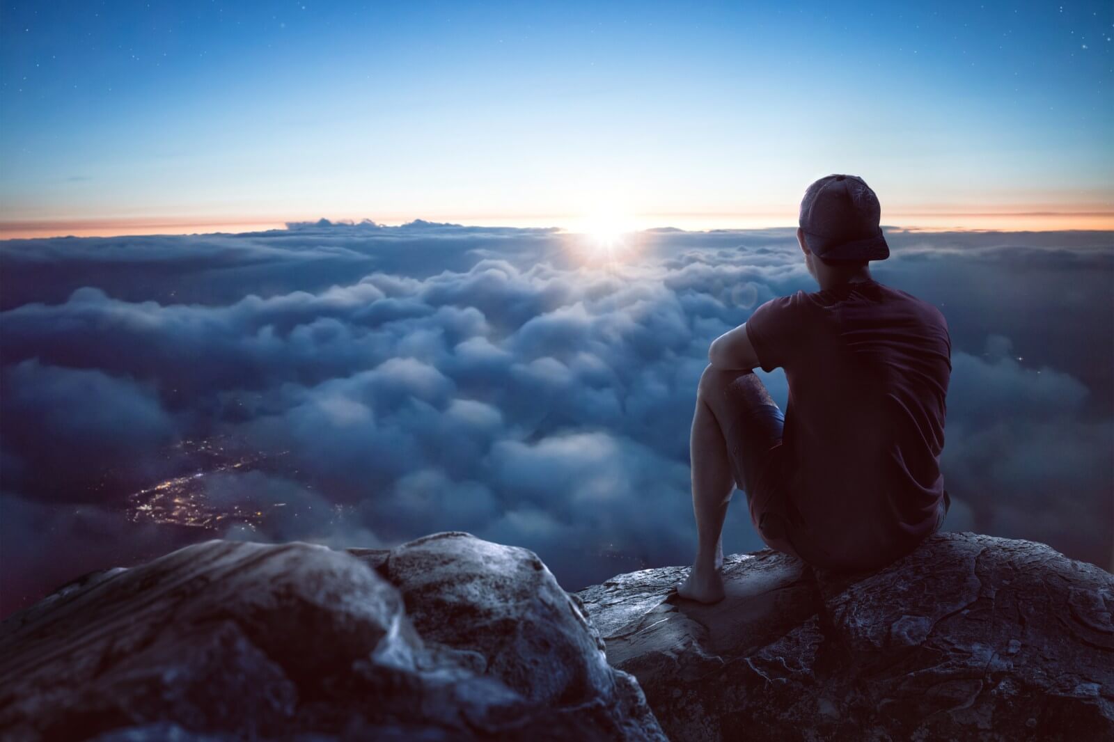 A young man is sitting on a rock on a summit overlooking a vast ocean of clouds. The sun is low and it is dark around him. Through the clouds you can see the lights of a city underneath.